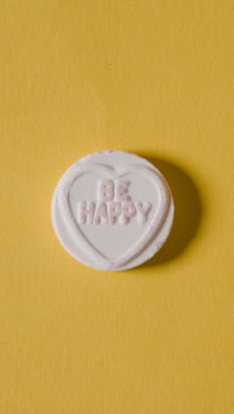 Vertical-Video-Of-Hand-Picking-Up-Heart-Candy-With-All-Yours-Message-On-Yellow-Background